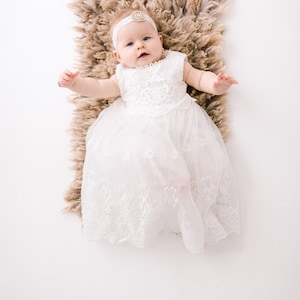 May include: A baby girl wearing a white lace dress and a headband with a jeweled embellishment lies on a brown fluffy rug.