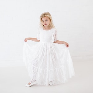 May include: A young girl wearing a white lace dress with a ruffled skirt and a gold leaf crown. She is standing on a white background.