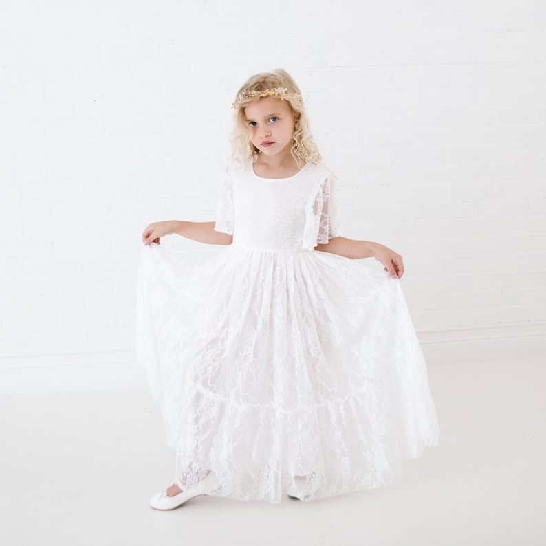 May include: A young girl wearing a white lace dress with a ruffled skirt and a gold leaf crown. She is standing on a white background.