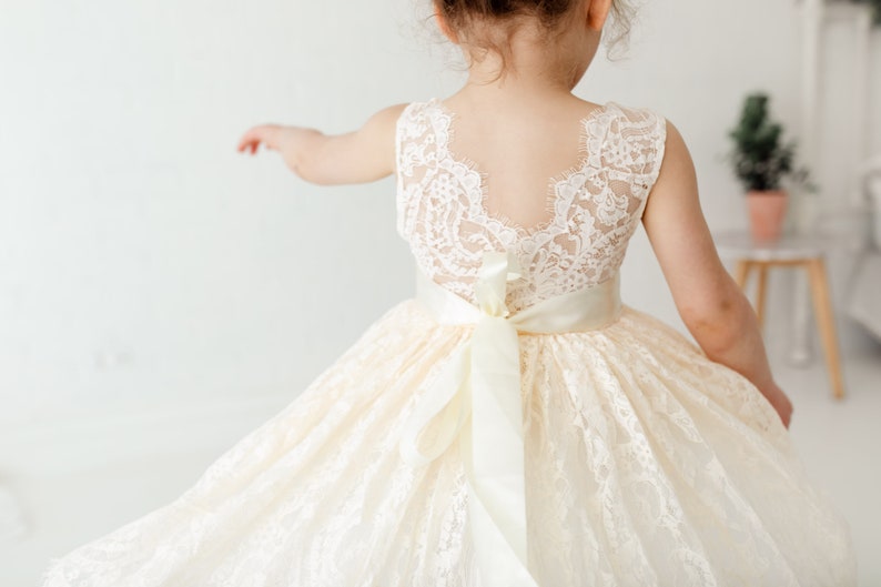 May include: A white lace dress with a satin ribbon sash. The dress has a scoop neckline and a sleeveless design. The back of the dress features a lace panel.