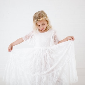 May include: A young girl wearing a white lace dress with a gold flower crown. The dress has a full skirt and short sleeves.