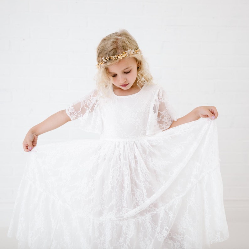 May include: A young girl wearing a white lace dress with a gold flower crown. The dress has a full skirt and short sleeves.