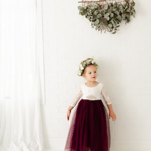 May include: A young girl wearing a white lace dress with a burgundy tulle skirt and a flower crown. She is standing in front of a white brick wall with a greenery wall hanging above her.