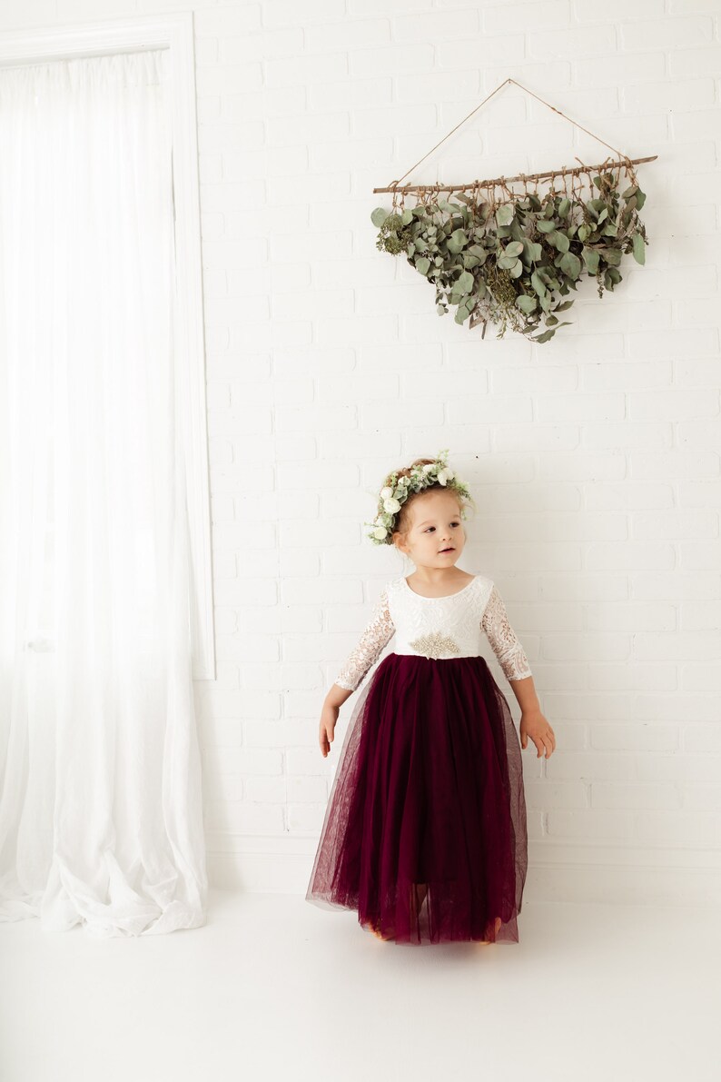 May include: A young girl wearing a white lace dress with a burgundy tulle skirt and a flower crown. She is standing in front of a white brick wall with a greenery wall hanging above her.