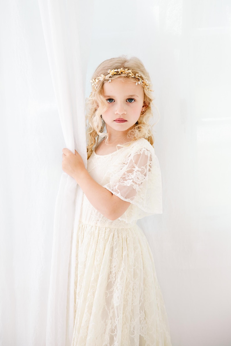 以下が含まれることがあります： 白いレースのドレスを着て、金色の花の冠をつけた若い女の子。彼女は白いカーテンの横に立ってカメラを見ている。