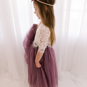 May include: A young girl wearing a white lace top with a mauve tulle skirt. She is wearing a burgundy velvet bow headband.