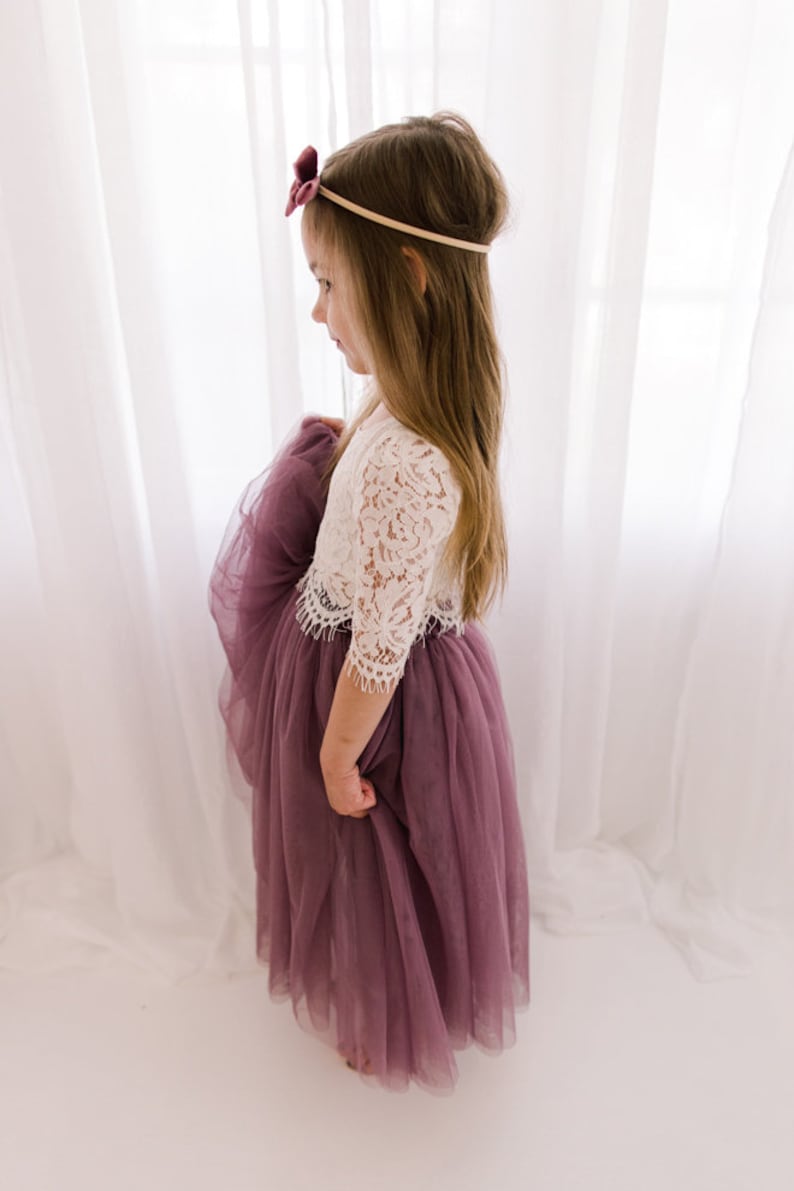 May include: A young girl wearing a white lace top with a mauve tulle skirt. She is wearing a burgundy velvet bow headband.