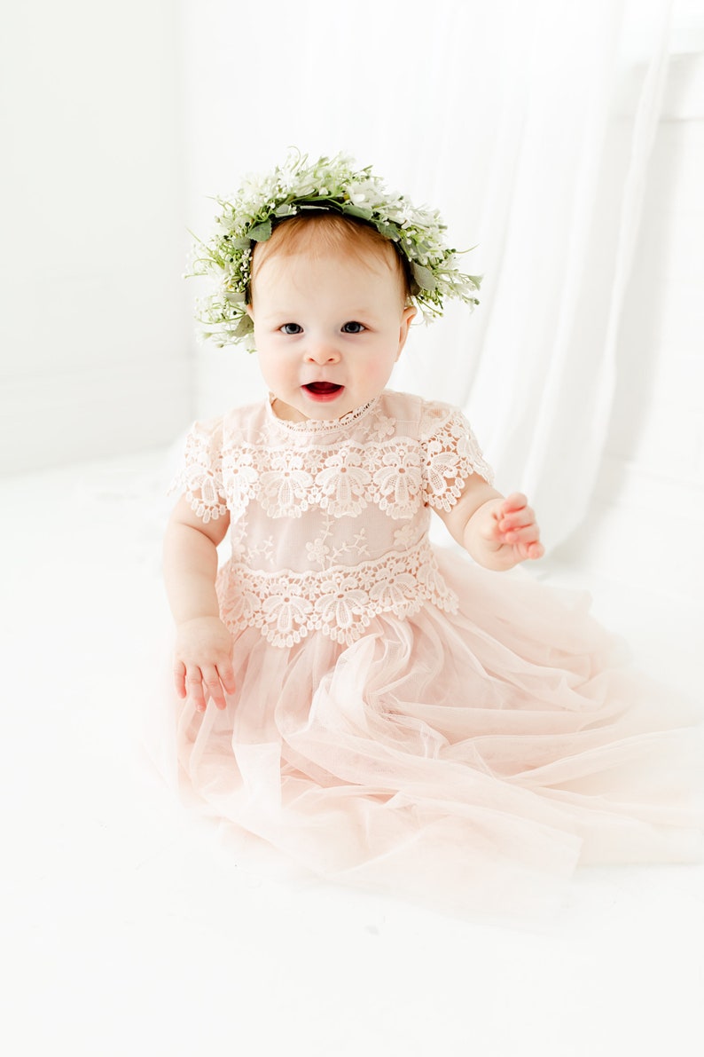 May include: A pale pink dress featuring a round neckline, short sleeves, and a bodice adorned with intricate white floral lace detailing. The skirt is constructed from layers of soft, sheer tulle fabric. A floral crown with white flowers and green leaves.