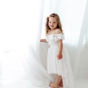 May include: A young girl with curly blonde hair wearing a white off-the-shoulder lace dress with a high-low hemline. She is standing in front of a white curtain and smiling at the camera.