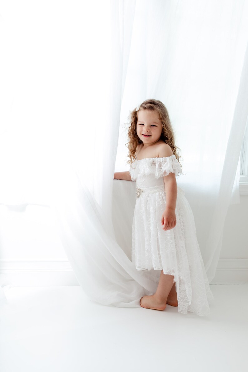 May include: A young girl with curly blonde hair wearing a white off-the-shoulder lace dress with a high-low hemline. She is standing in front of a white curtain and smiling at the camera.
