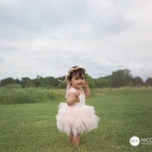 May include: A young girl wearing a pink tulle dress and a flower crown stands in a grassy field. She is looking to the side and smiling.