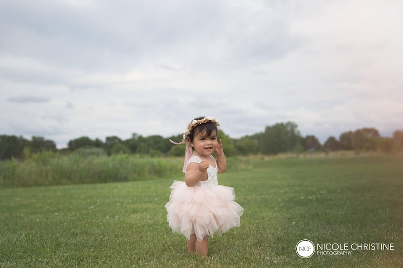 May include: A young girl wearing a pink tulle dress and a flower crown stands in a grassy field. She is looking to the side and smiling.