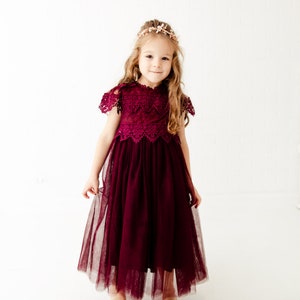 May include: A young girl wearing a burgundy tulle dress with a lace bodice and a floral headband. The dress has a scalloped hemline and the girl is standing in front of a white background.