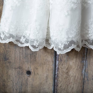 May include: A white dress featuring an intricate floral lace overlay with a scalloped hemline. The delicate embroidery covers the sheer fabric, which is layered over a solid white base, creating a textured and detailed appearance throughout.