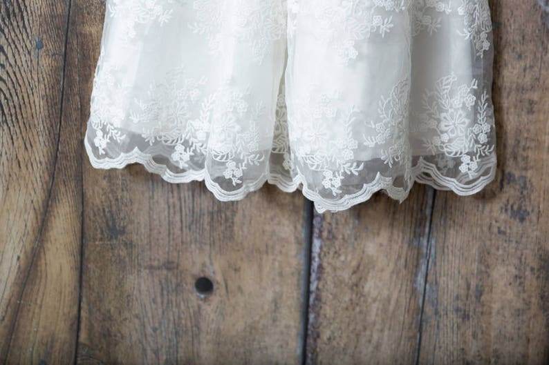May include: A white dress featuring an intricate floral lace overlay with a scalloped hemline. The delicate embroidery covers the sheer fabric, which is layered over a solid white base, creating a textured and detailed appearance throughout.