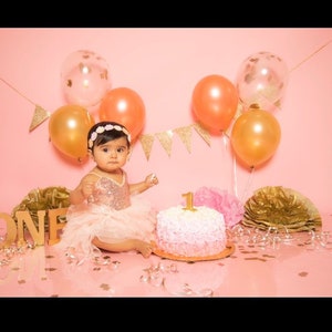 May include: A baby girl wearing a pink sequined dress and a flower crown sits in front of a pink cake decorated with a gold number one. The cake is surrounded by gold and pink balloons and confetti.