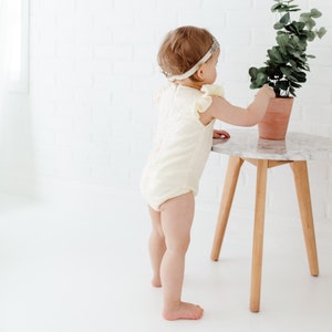 Puede incluir: Una ni&ntilde;a peque&ntilde;a con un mono blanco de lentejuelas y volantes y una diadema brillante se encuentra junto a una peque&ntilde;a mesa de madera con una planta en maceta.