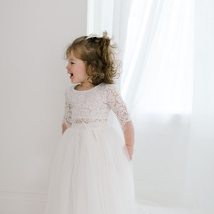 May include: A young girl wearing a white lace dress with a tulle skirt. She is standing in front of a white window with white curtains.