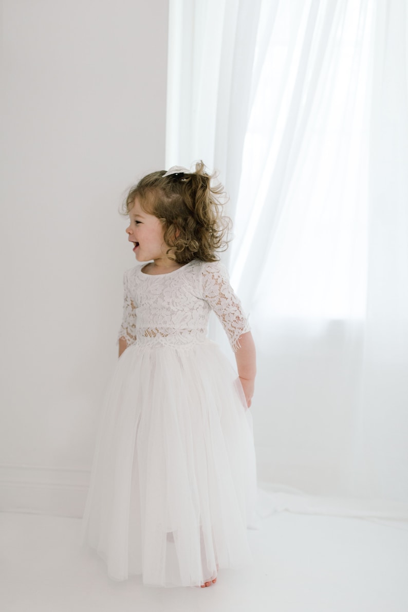 May include: A young girl wearing a white lace dress with a tulle skirt. She is standing in front of a white window with white curtains.