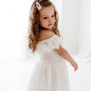 May include: A young girl with curly hair wears a white off-the-shoulder lace dress. She has a pearl hair clip in her hair.