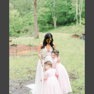 May include: A white sleeveless wedding dress with a lace bodice and a full tulle skirt, paired with two matching pale pink flower girl dresses featuring lace bodices and short sleeves, held together in a group embrace with a bouquet of white flowers.