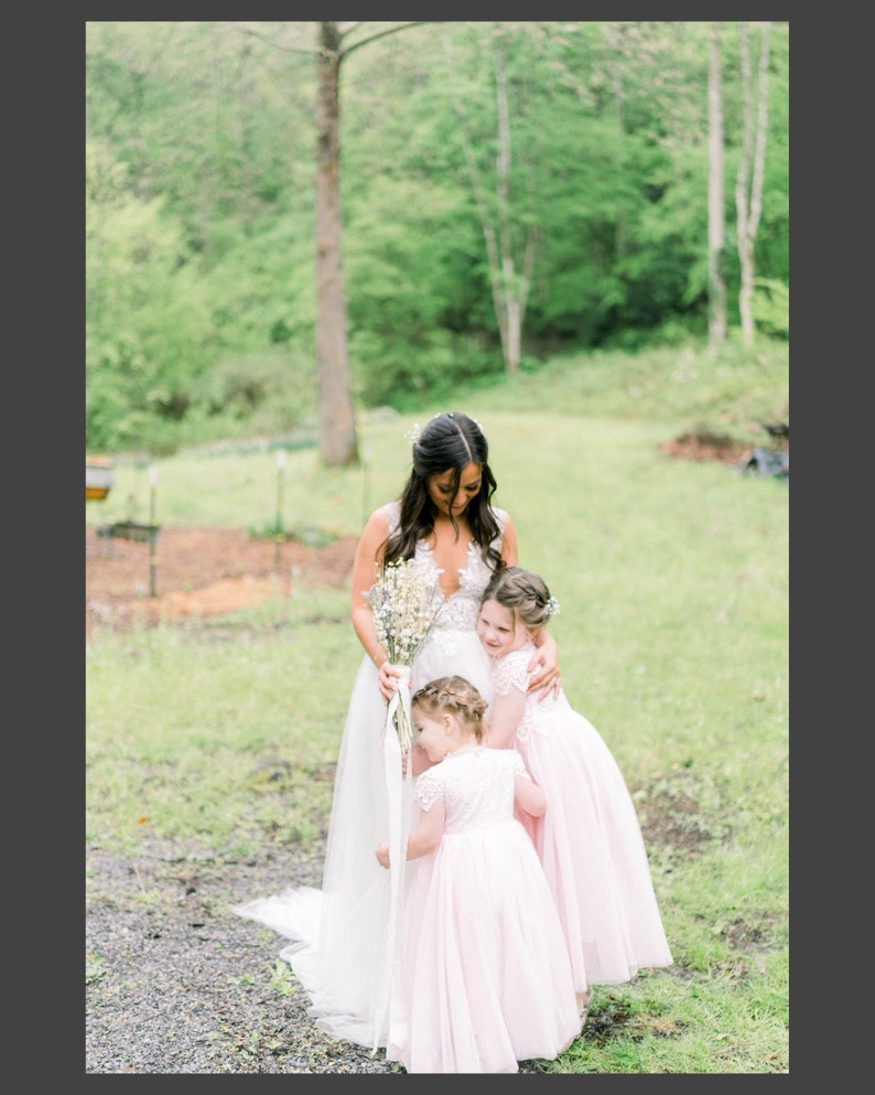 May include: A white sleeveless wedding dress with a lace bodice and a full tulle skirt, paired with two matching pale pink flower girl dresses featuring lace bodices and short sleeves, held together in a group embrace with a bouquet of white flowers.