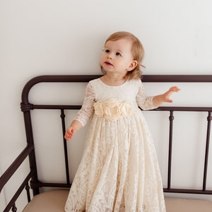 May include: A young girl wearing a white lace dress with a satin flower sash. The dress has long sleeves and a full skirt. The girl is standing on a brown couch.