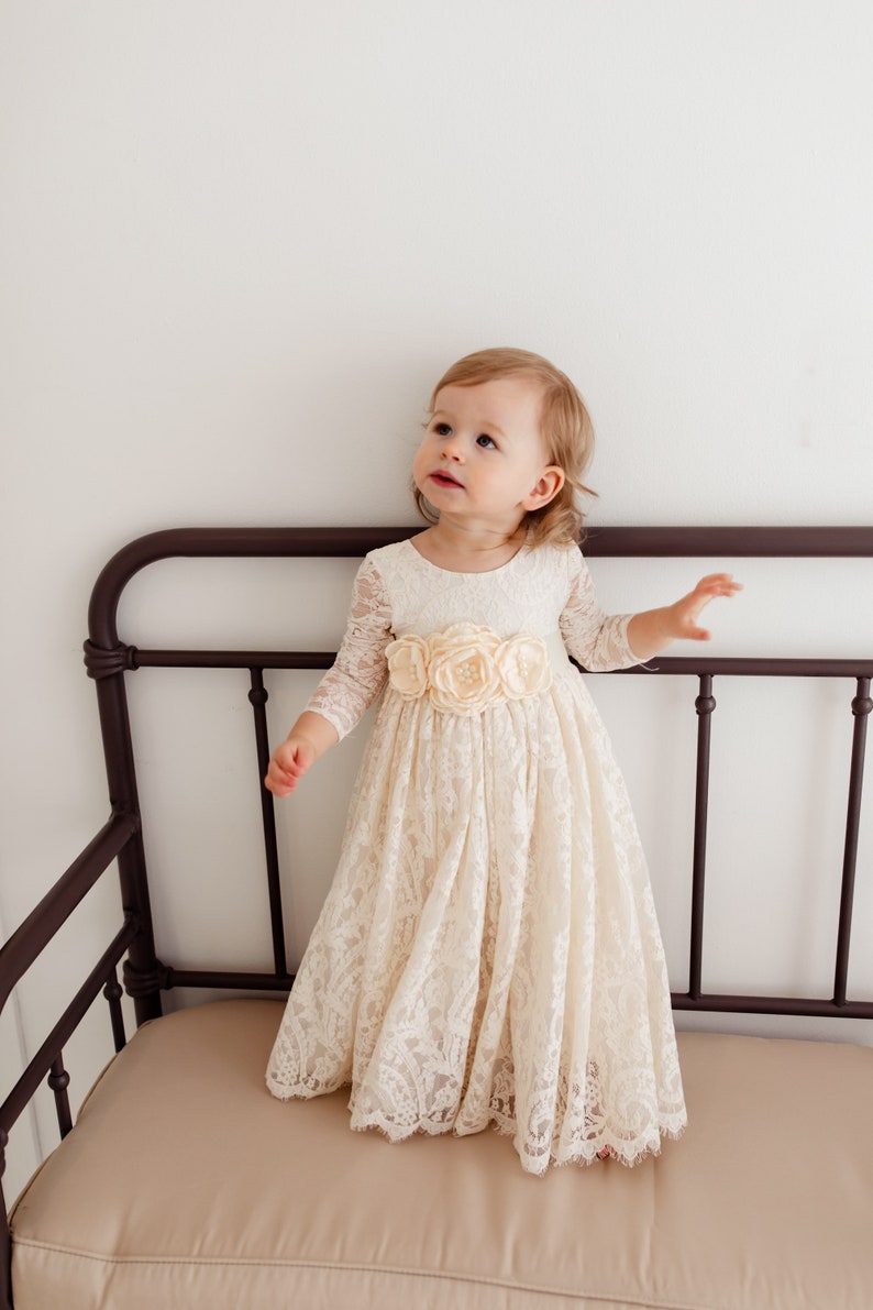 May include: A young girl wearing a white lace dress with a satin flower sash. The dress has long sleeves and a full skirt. The girl is standing on a brown couch.