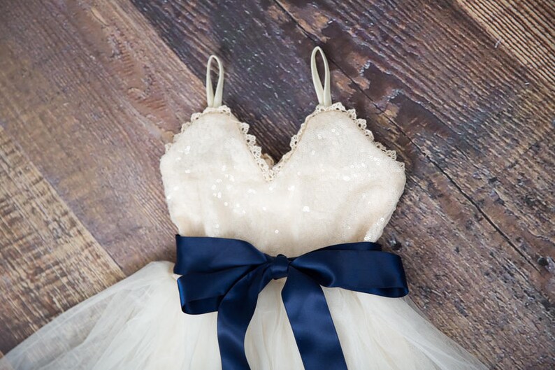 May include: A white tulle dress with a sequined bodice and a navy blue satin ribbon tied around the waist.