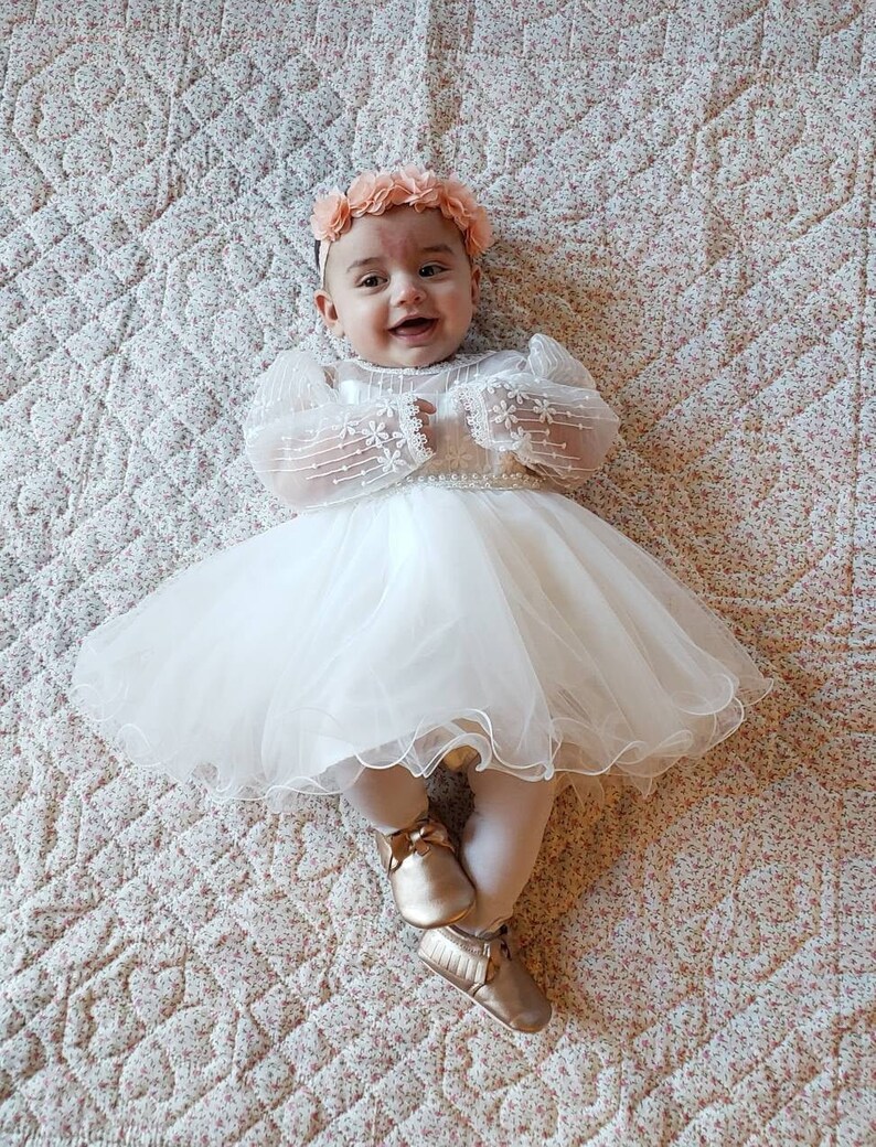 K&ouml;nnte beinhalten: Ein kleines M&auml;dchen in einem wei&szlig;en T&uuml;llkleid mit einem Blumenhaarband und goldenen Schuhen. Das Kleid hat Spitzen&auml;rmel und einen Perlenbesatz.