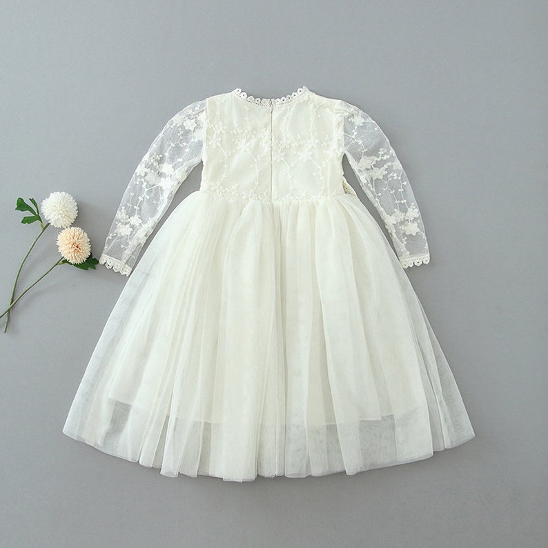 May include: A cream-colored dress featuring long, sheer lace sleeves with floral embroidery, a round neckline with scalloped trim, and a full, layered tulle skirt. The bodice is fitted, and the back includes a central zipper closure.