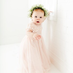 May include: A pale pink dress featuring a lace bodice with short sleeves and a layered tulle skirt. The garment is paired with a floral crown made of small white flowers and green leaves, worn on the head to complement the soft color palette.