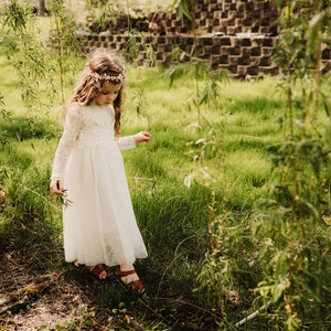May include: A young girl wearing a white dress and a flower crown walks through a grassy field.