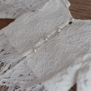 May include: A white lace garment featuring a floral pattern and a vertical row of four small, round, white pearl-like buttons. The fabric is semi-sheer with intricate openwork detailing throughout the material, showcasing a delicate texture.