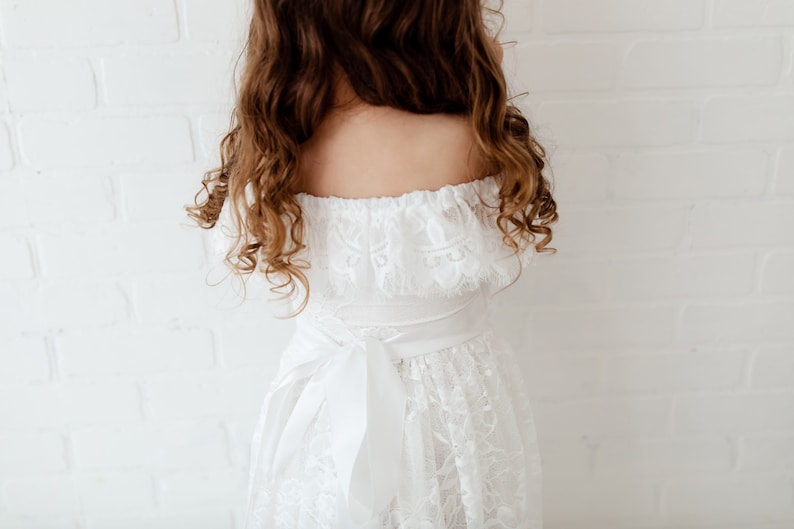 May include: White off-the-shoulder dress with lace detailing and a white satin ribbon tied at the waist.
