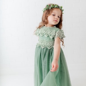 May include: A young girl wearing a sage green tulle dress with lace detailing and a leafy green crown. The dress has a scalloped lace trim and a full skirt.