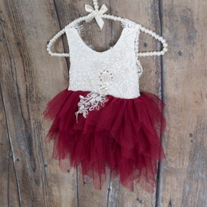 Puede incluir: Vestido de beb&eacute; o ni&ntilde;a peque&ntilde;a, de encaje blanco y tul burdeos, colgado de una percha de perlas. El corpi&ntilde;o es de encaje blanco con adornos florales, y la falda es de tul burdeos en capas.
