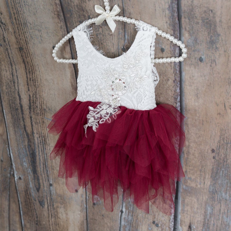 Puede incluir: Vestido de beb&eacute; o ni&ntilde;a peque&ntilde;a, de encaje blanco y tul burdeos, colgado de una percha de perlas. El corpi&ntilde;o es de encaje blanco con adornos florales, y la falda es de tul burdeos en capas.