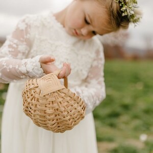 May include: A young girl in a white lace dress holds a woven basket. She is wearing a flower crown and looking down at the basket.