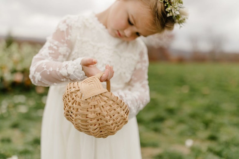 May include: A young girl in a white lace dress holds a woven basket. She is wearing a flower crown and looking down at the basket.