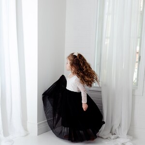 May include: A long-sleeved dress featuring a white lace bodice with a scoop neckline and a voluminous, floor-length black tulle skirt. The garment is designed with a fitted top that transitions into a full, layered skirt, creating a distinct contrast between the white lace and the solid black fabric.
