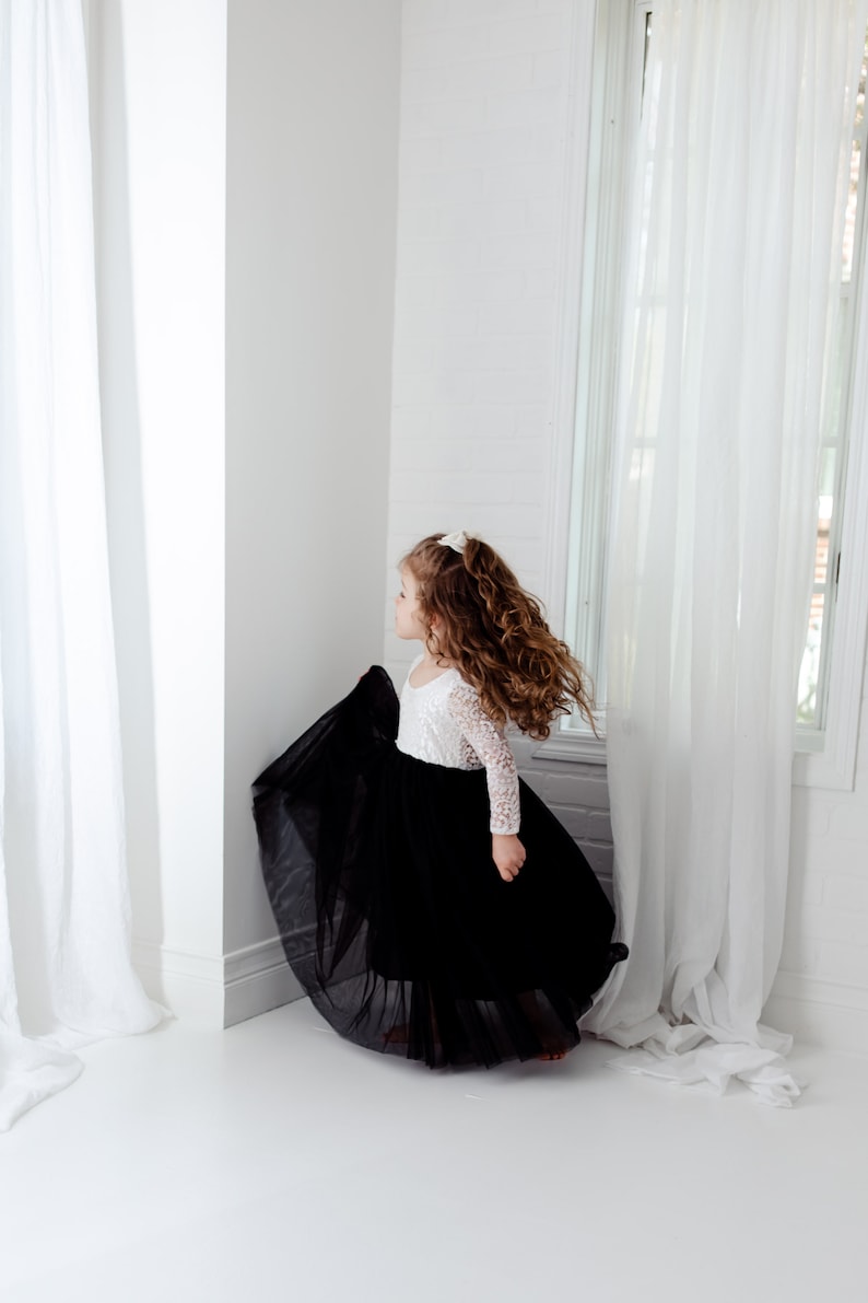 May include: A long-sleeved dress featuring a white lace bodice with a scoop neckline and a voluminous, floor-length black tulle skirt. The garment is designed with a fitted top that transitions into a full, layered skirt, creating a distinct contrast between the white lace and the solid black fabric.