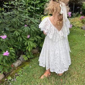 May include: A young girl wearing a white lace dress with a ruffled neckline and a flower crown. She is standing in a garden with green grass and flowers.