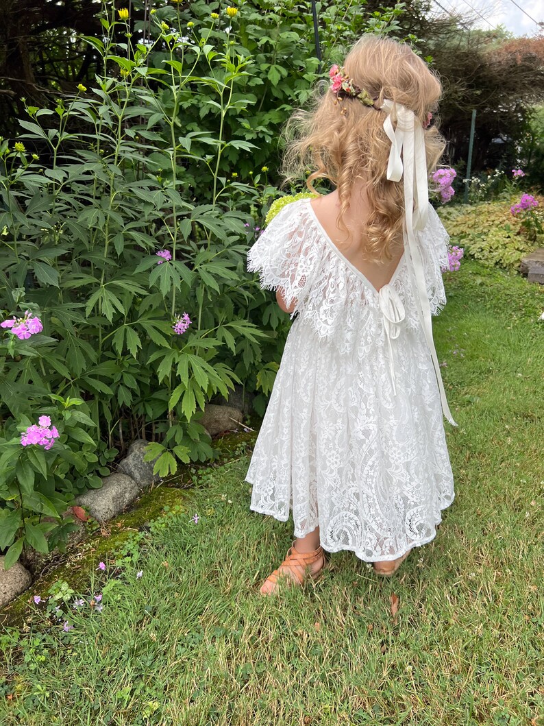 May include: A young girl wearing a white lace dress with a ruffled neckline and a flower crown. She is standing in a garden with green grass and flowers.