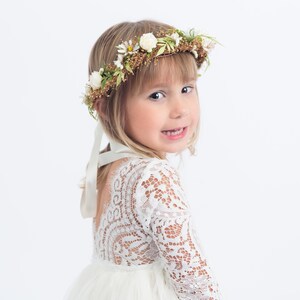 Pu&ograve; includere: Una bambina che indossa un abito bianco di pizzo a maniche lunghe e una corona di fiori. La corona &egrave; fatta di fiori bianchi e foglie verdi.