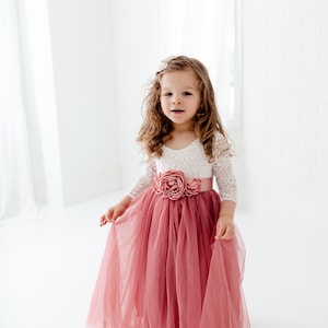 May include: A young girl wearing a white lace top with a pink flower on the bodice and a long, flowing pink tulle skirt.