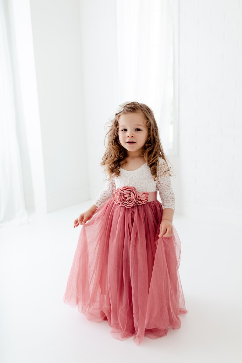 May include: A young girl wearing a white lace top with a pink flower on the bodice and a long, flowing pink tulle skirt.