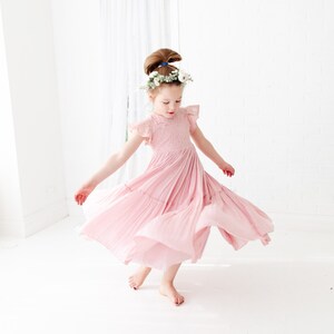 May include: A young girl wearing a pink, tiered, lace dress with a floral crown. She is twirling and smiling.