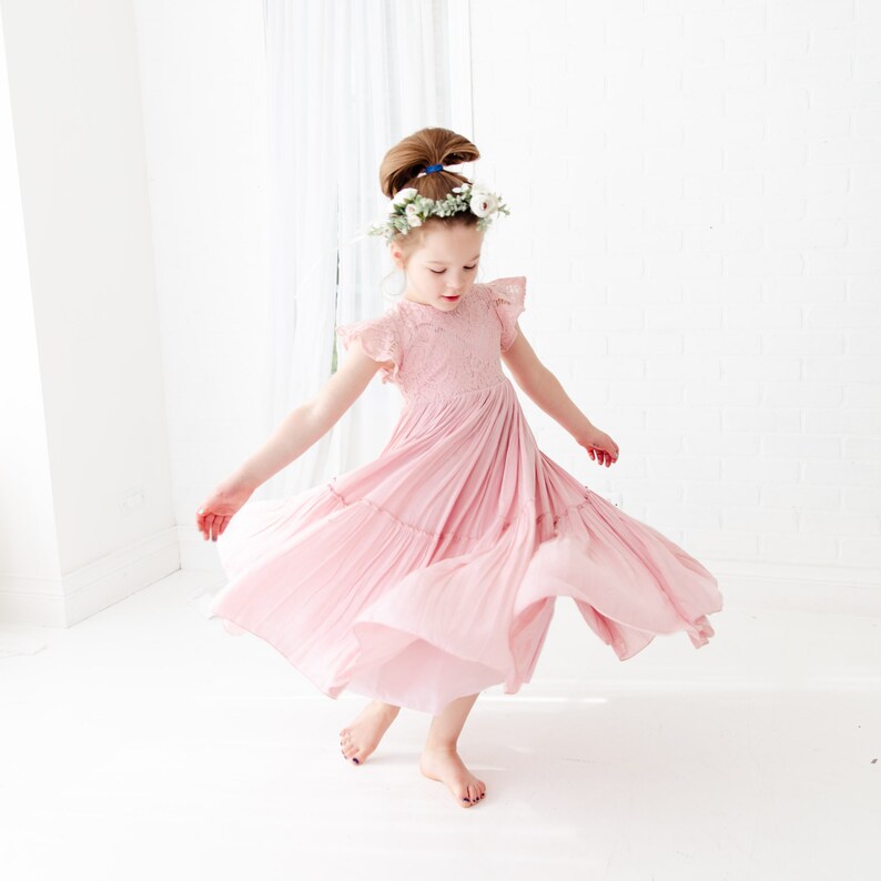 May include: A young girl wearing a pink, tiered, lace dress with a floral crown. She is twirling and smiling.