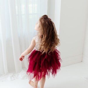 Puede incluir: Una ni&ntilde;a con un top de encaje blanco y una falda de tul rojo oscuro en capas. La ni&ntilde;a mira hacia arriba, con cabello casta&ntilde;o largo y suelto. El vestido contrasta vivamente con el fondo blanco.